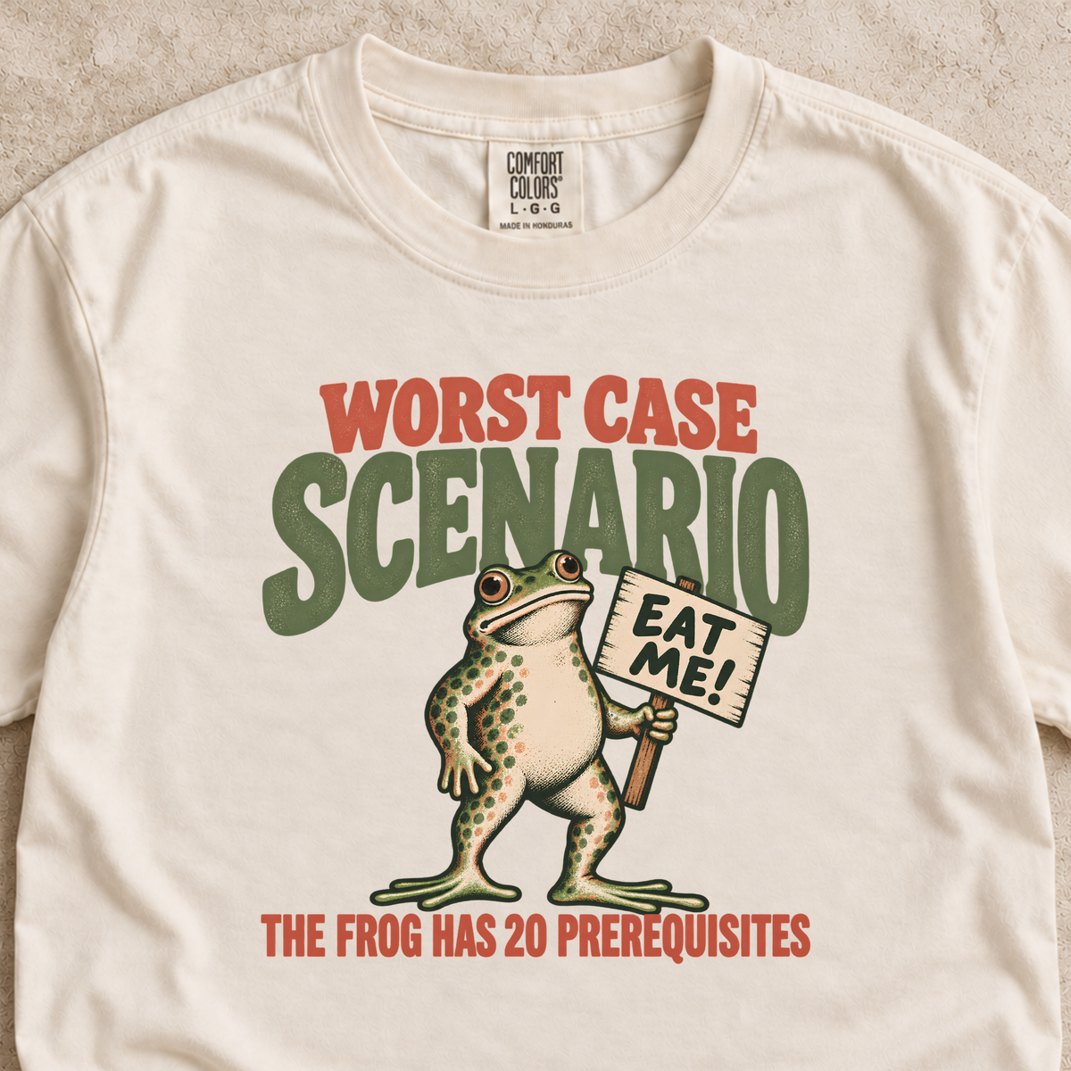 An off-white crewneck t-shirt featuring a retro-style graphic of a frog holding a sign that says 'EAT ME!'. The text reads 'WORST CASE SCENARIO' in red and green, and 'THE FROG HAS 20 PREREQUISITES' in red at the bottom.